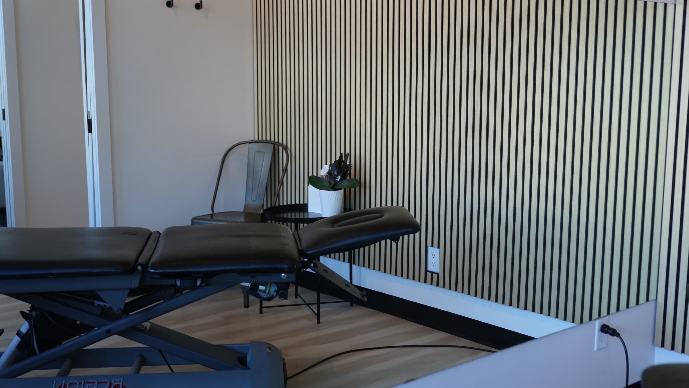 Treatment room with a black padded adjustable treatment table in the foreground, a metal chair, a round side table with a potted white planter and plant, and a wall of vertical light wood slat panels