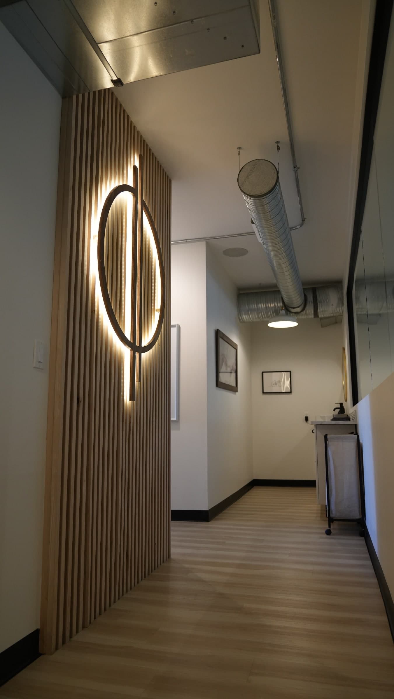 Hallway in a clinic with a backlit circular wooden wall feature over vertical timber slats, a stainless ventilation duct overhead, framed artwork, and a sink with a laundry hamper beside it