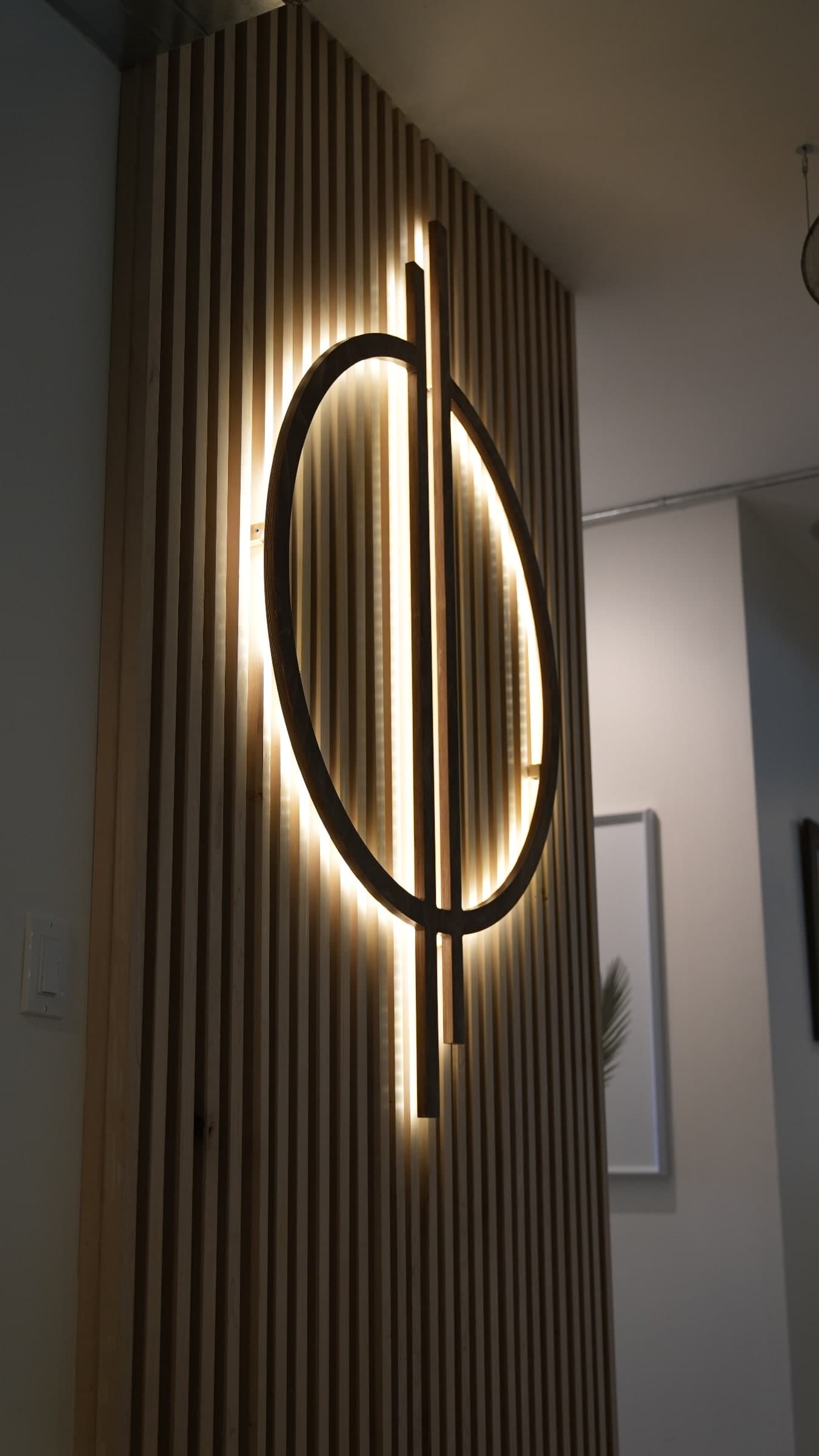 Backlit circular wooden wall feature mounted over vertical wood slat panels with warm LED lighting, in an interior hallway