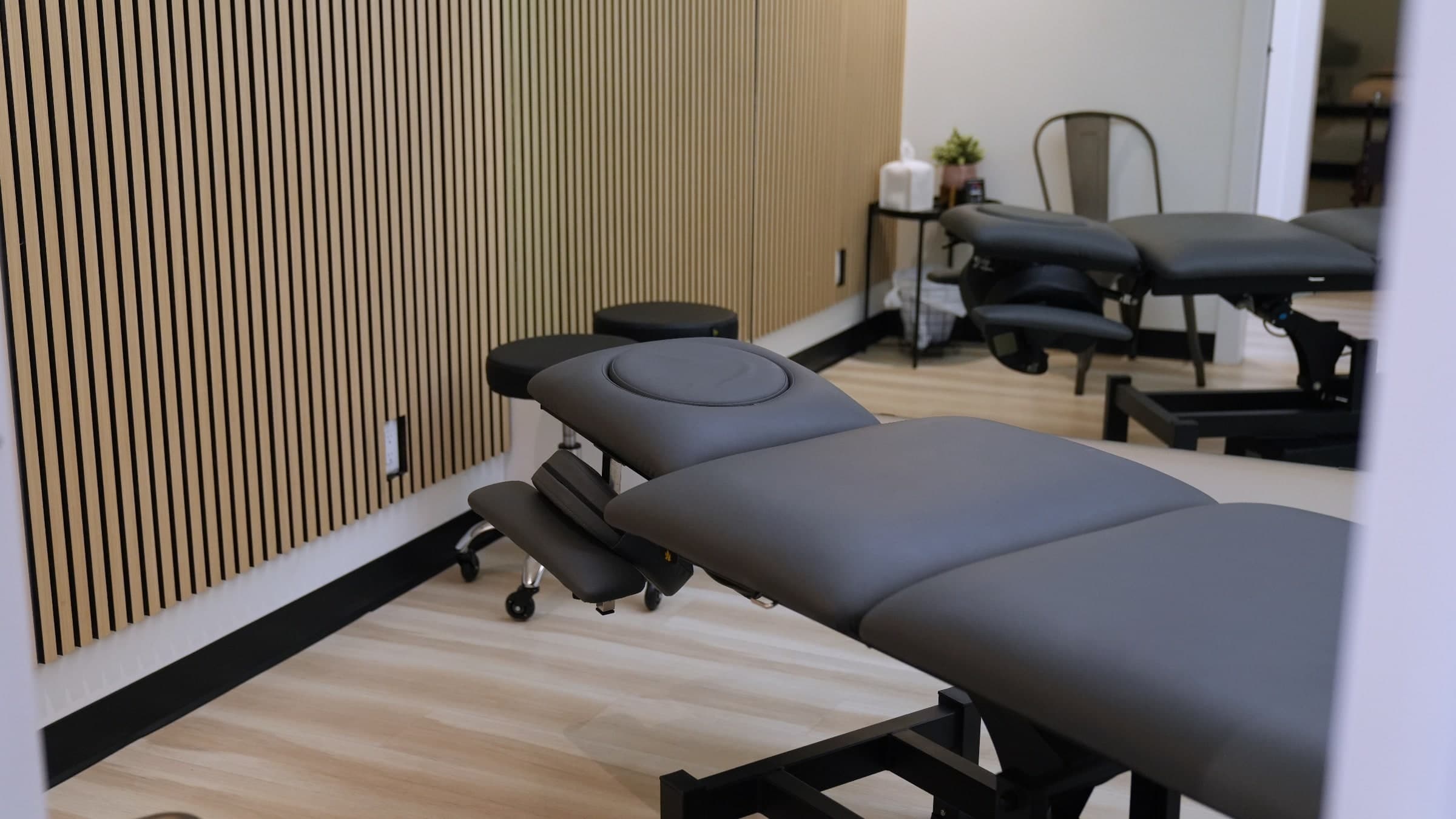 Treatment room with grey padded chiropractic adjusting tables, a black stool, wood slat accent wall and a potted plant in Calgary