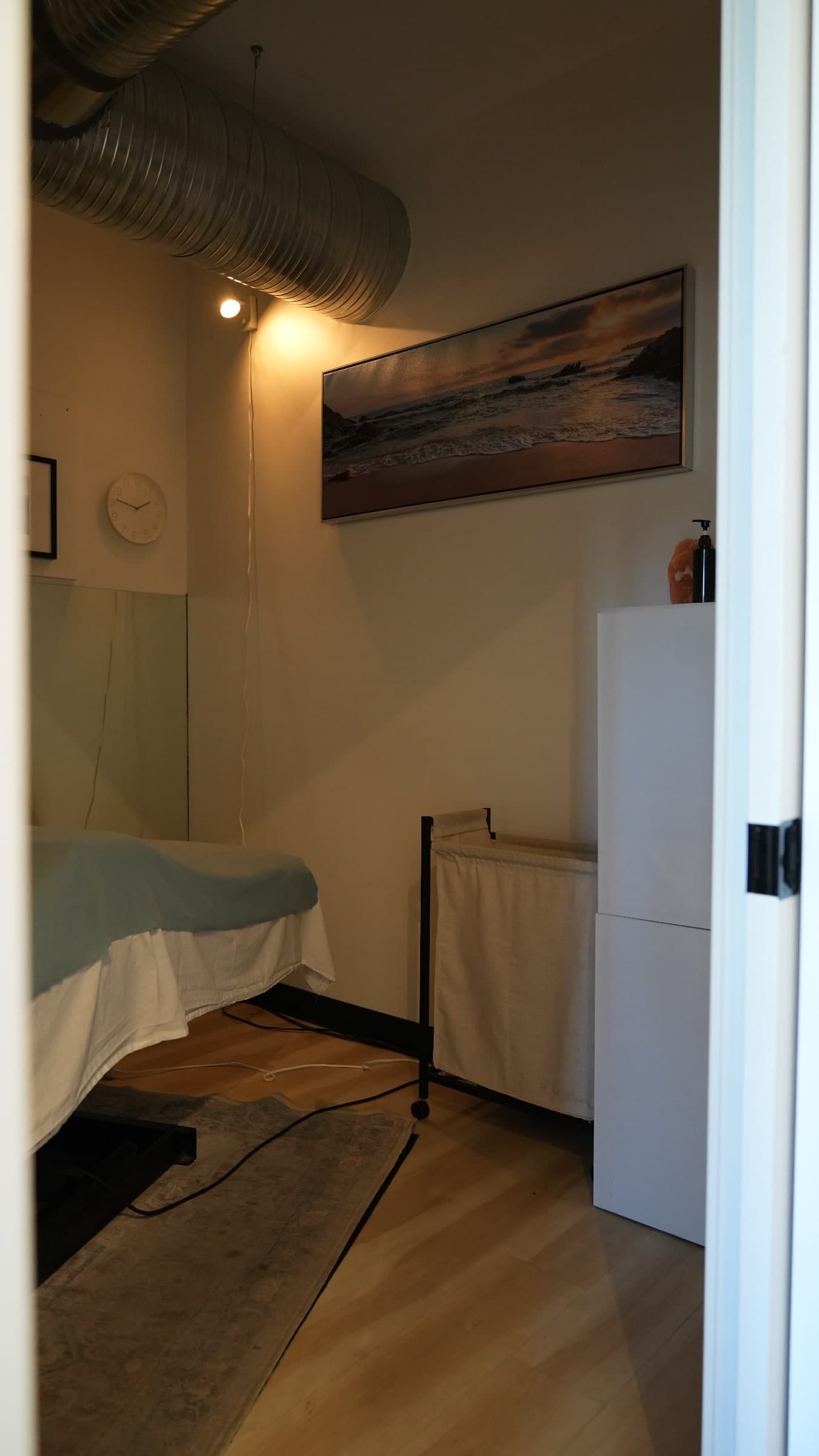 Treatment room with a massage table draped in white and pale green linens, a framed coastal sunset photograph on the wall, an exposed ventilation duct on the ceiling, and a wall-mounted spotlight, Calgary