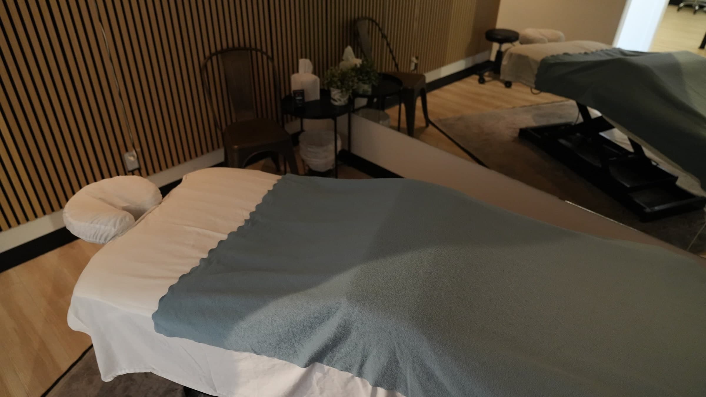 Two massage treatment beds with white fitted sheets and grey blankets, a cream face cradle on the left bed, beside a wooden slatted wall, a small side table with tissues and a potted plant, and a rolling stool