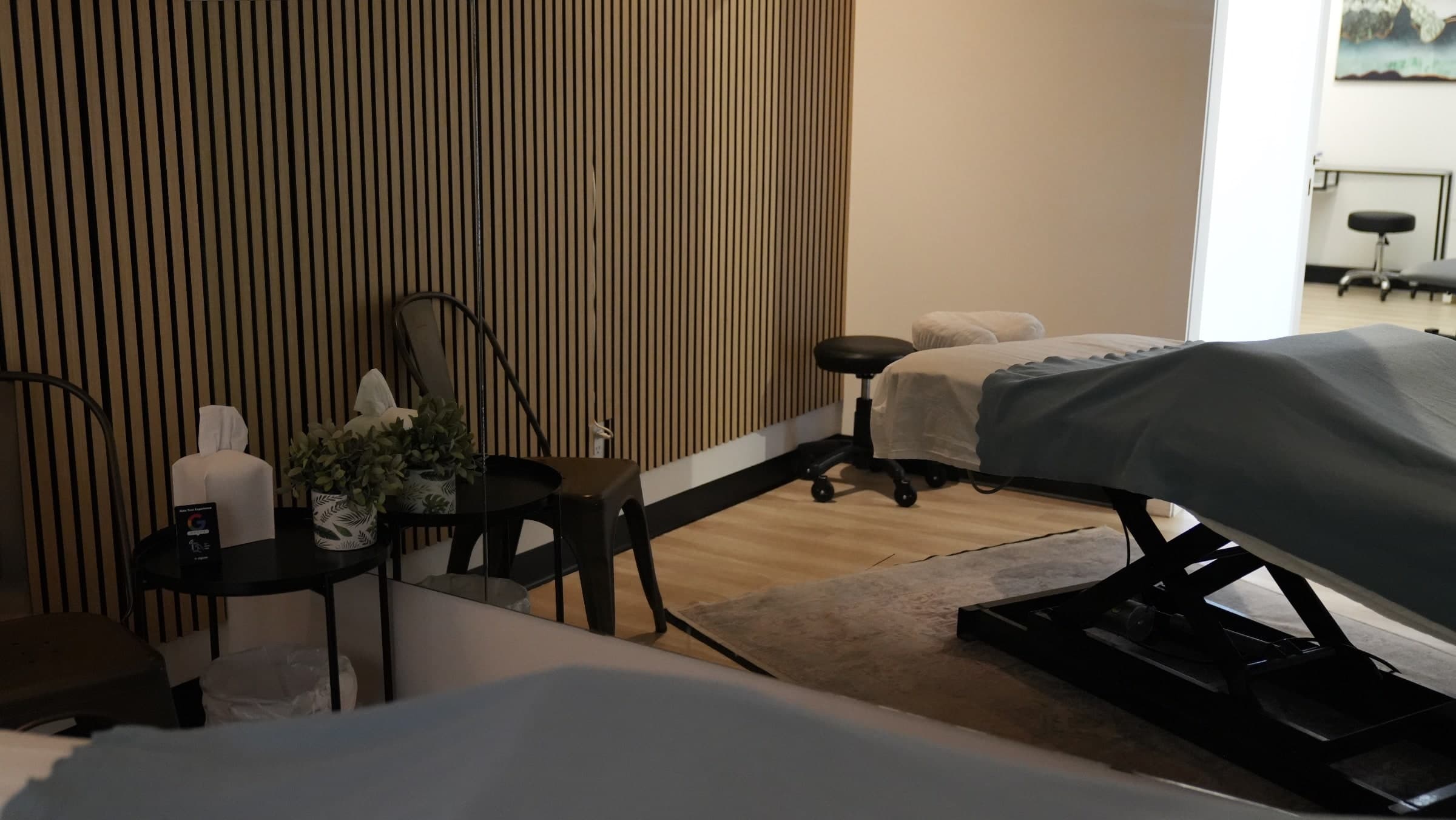 Treatment room with two massage tables, grey linens, a black rolling stool, potted plants, tissue boxes and wood slat accent wall