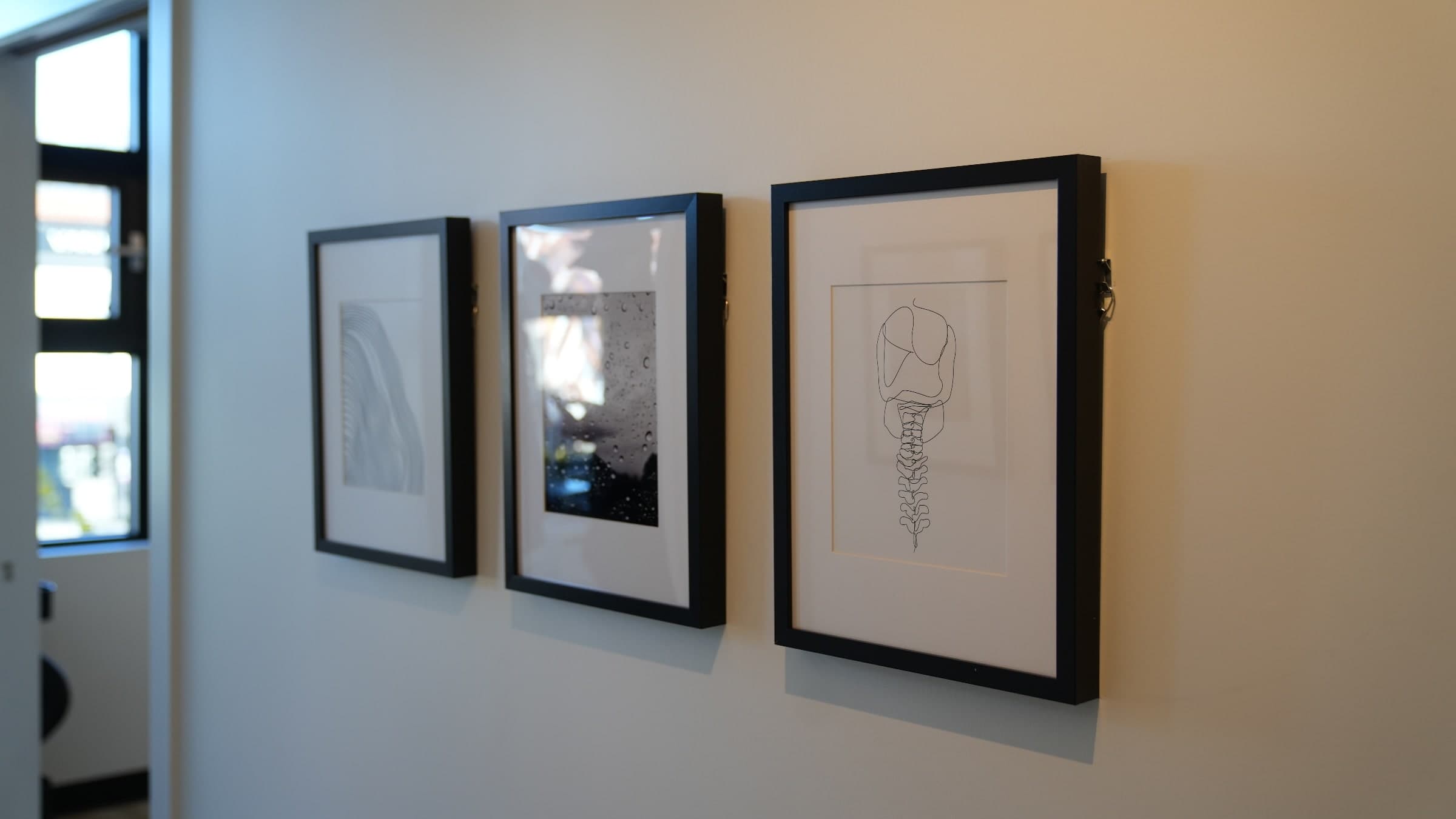 Three framed black-and-white artworks on a wall: a fine line-pattern curve on the left, a close-up photo of water droplets on glass in the centre, and a continuous line drawing of a spine on the right