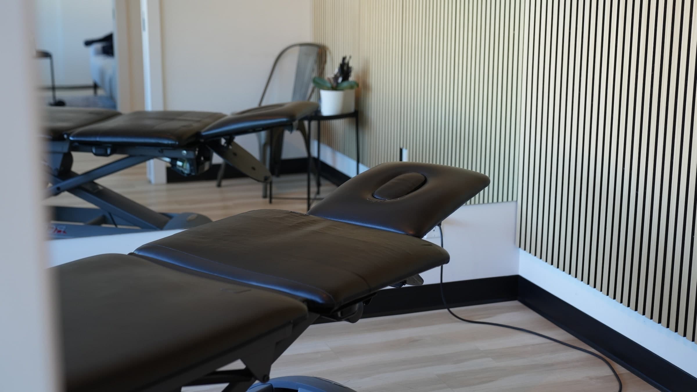 Treatment room with two black padded treatment tables with adjustable headrests, slatted wood wall panels, a potted plant on a side table, and light wood flooring