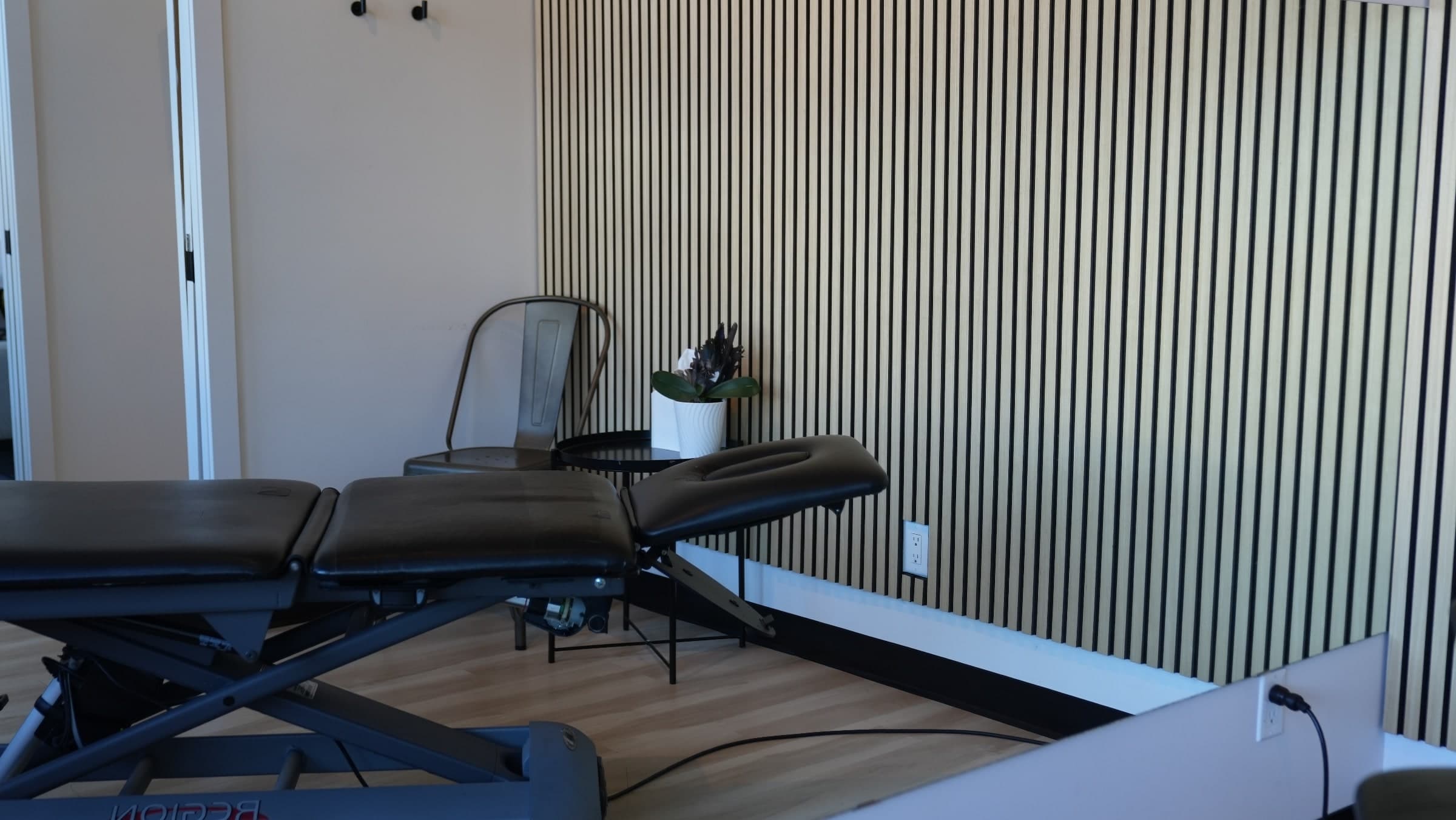 Treatment room with a black adjustable treatment table, a metal chair, a small side table with a potted plant, and a slatted wood-panel accent wall, Calgary