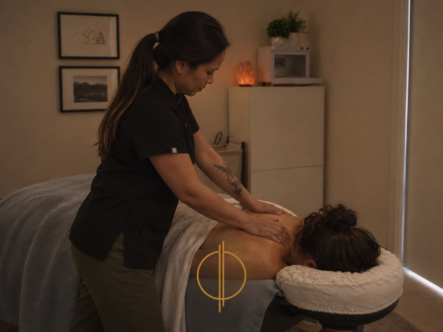 A massage therapist in a black top and olive pants working on the upper back of a patient lying face down on a massage table in a warmly lit treatment room with a salt lamp and framed prints in Calgary