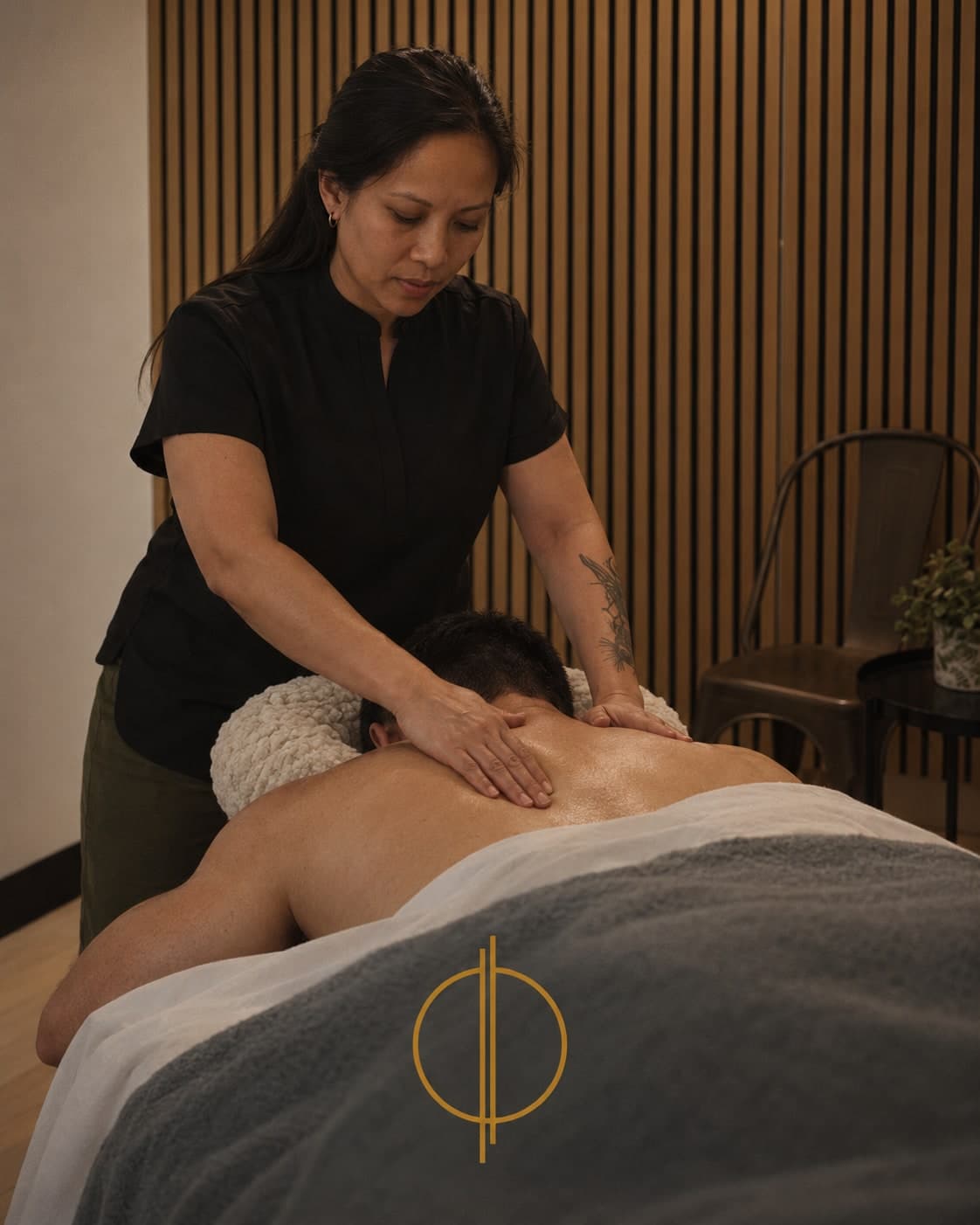 Massage therapist applying hands to the upper back of a shirtless patient lying face down on a massage table in a warm treatment room with wood slat walls