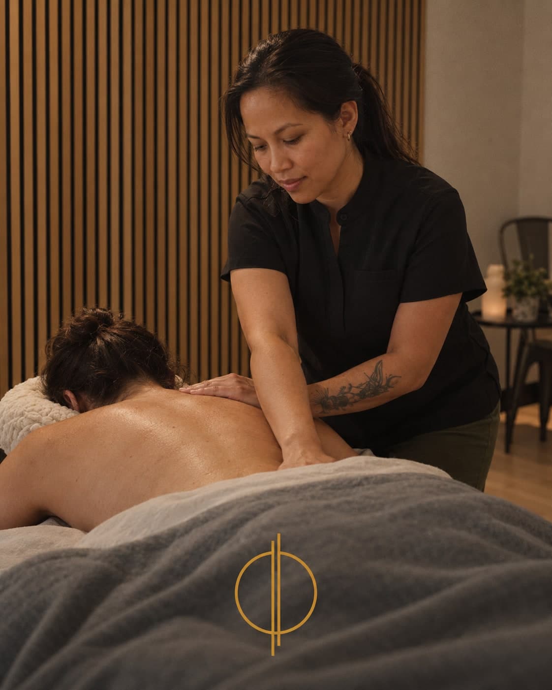 Massage therapist in a black uniform working on the upper back of a patient lying face down on a massage table in a warm wood-paneled treatment room in Calgary