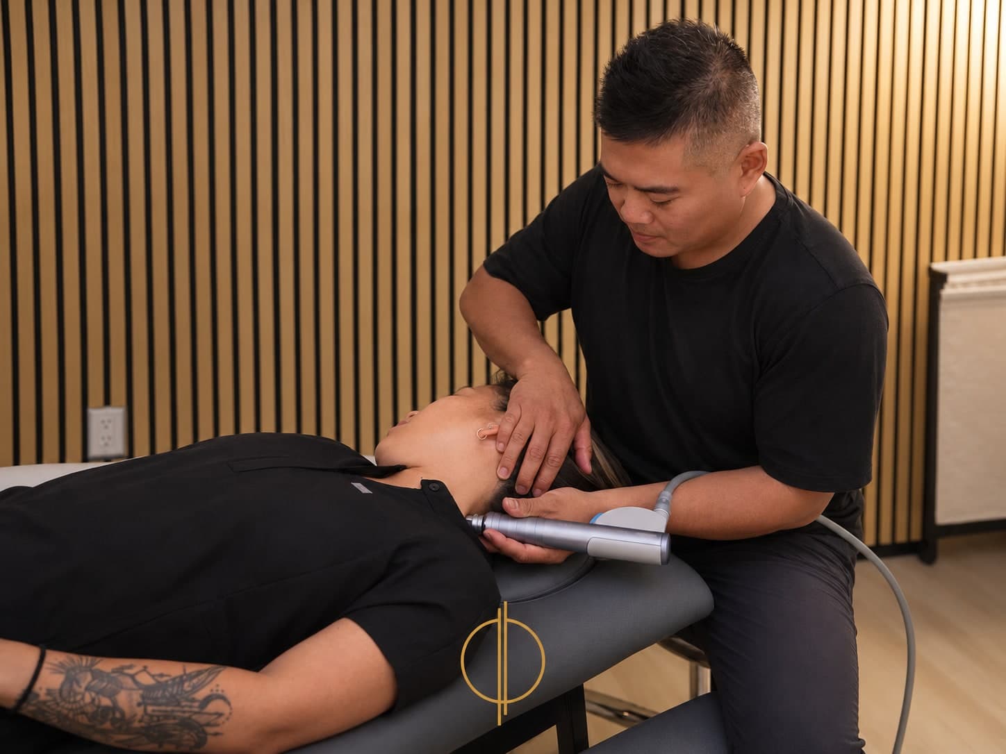 Male clinician in a black t-shirt treating a patient lying face up on a treatment table, holding a handheld percussion device to the patient's neck while supporting the head with one hand, wood-slat wall behind, Calgary