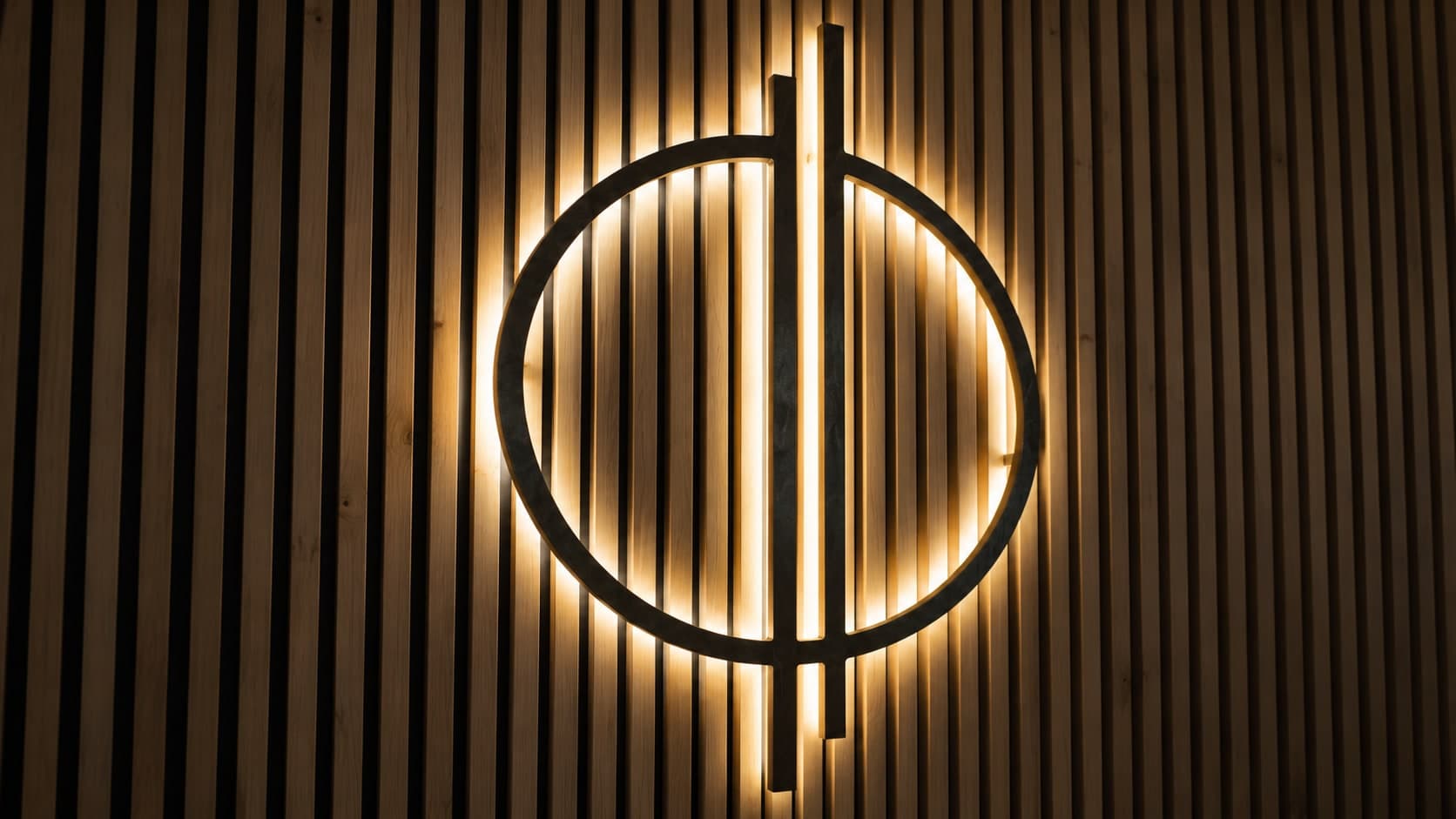 Illuminated black circular logo with a vertical bar through its center, mounted on backlit wood slat wall, Calgary
