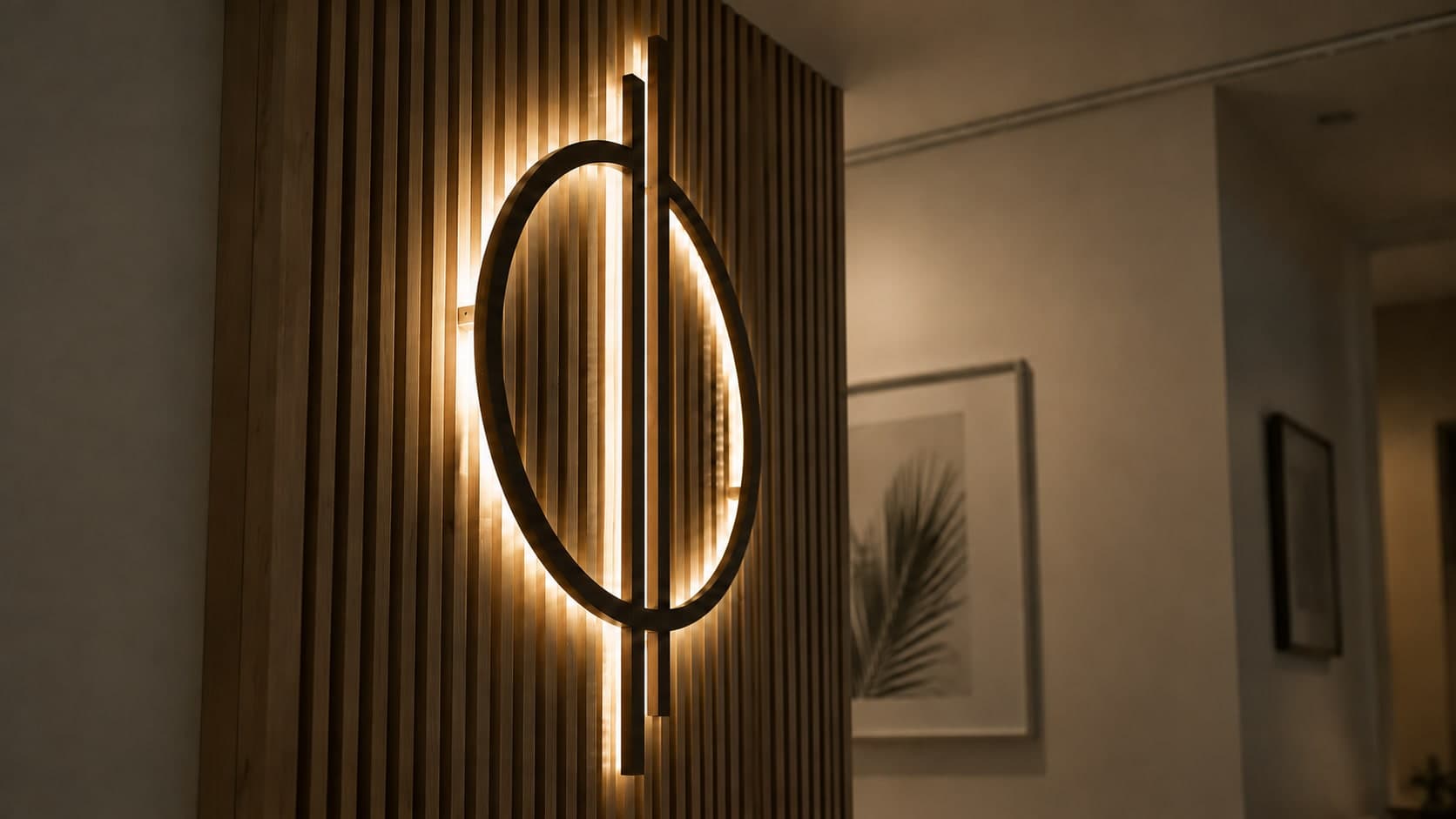 Warm-lit interior wall with vertical wood slat panelling and a backlit oval wooden wall sculpture, a framed black-and-white palm leaf print beside it
